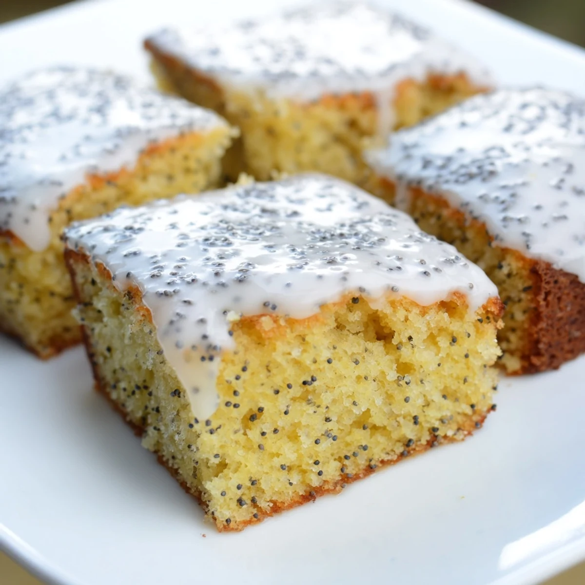 A slice of moist Lemon Drizzle Traybake with Poppy Seeds on a white plate with a fork, ideal for afternoon tea.