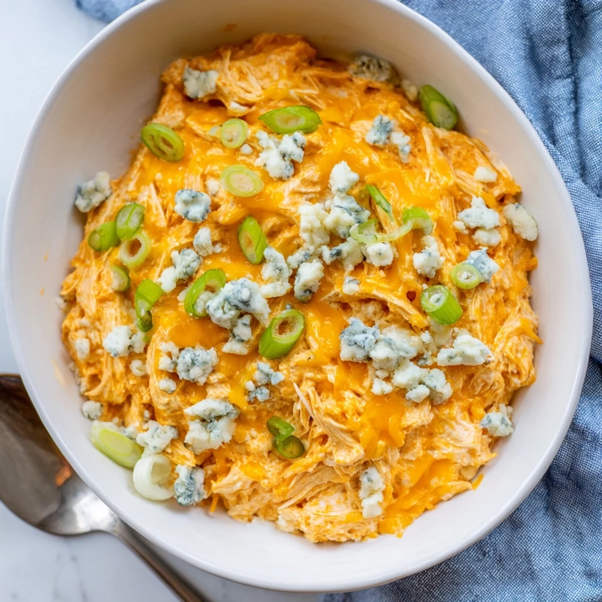 A close-up of golden, melted cheddar topping on the rich, orange-hued Touchdown Buffalo Chicken Dip, with blue cheese crumbles and a serving platter nearby.  