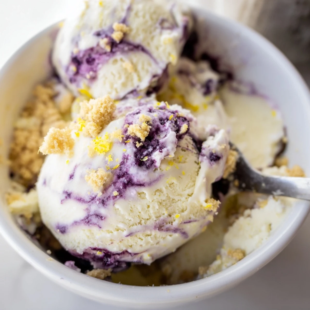 A close-up view of Lemon Blueberry Cheesecake Ice Cream in a chilled glass bowl, garnished with fresh lemon zest.
